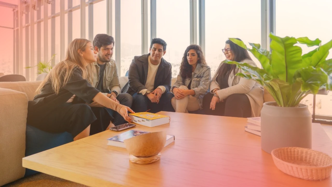 Grupo de jovens profissionais participando de uma reunião de equipe em um ambiente de escritório moderno, com muita luz natural e plantas decorativas.