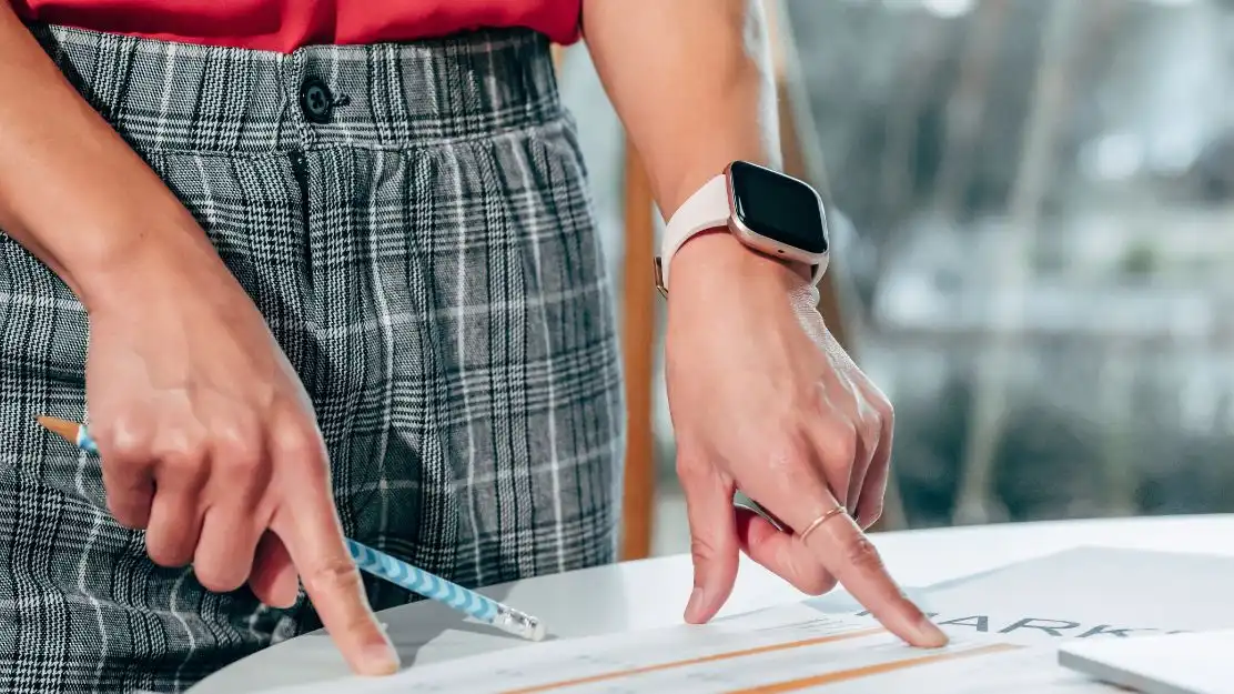 Mulher apontando para documentos sobre uma mesa, com uma caneta nas mãos e um relógio inteligente no pulso. A imagem reflete um ambiente de trabalho educacional e produtivo.