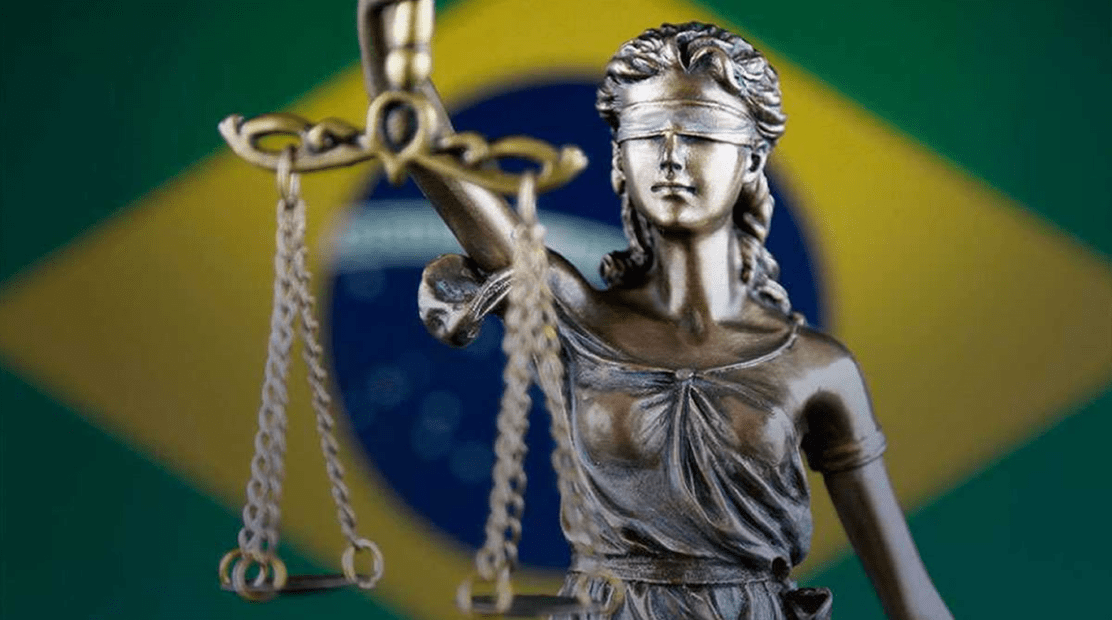imagem mostra a bandeira do brasil ao fundo, desfocada e colorida, com a estátua dourada a frente de uma mulher com olhos vendados segurando uma balança, conhecia como Dama da Justiça