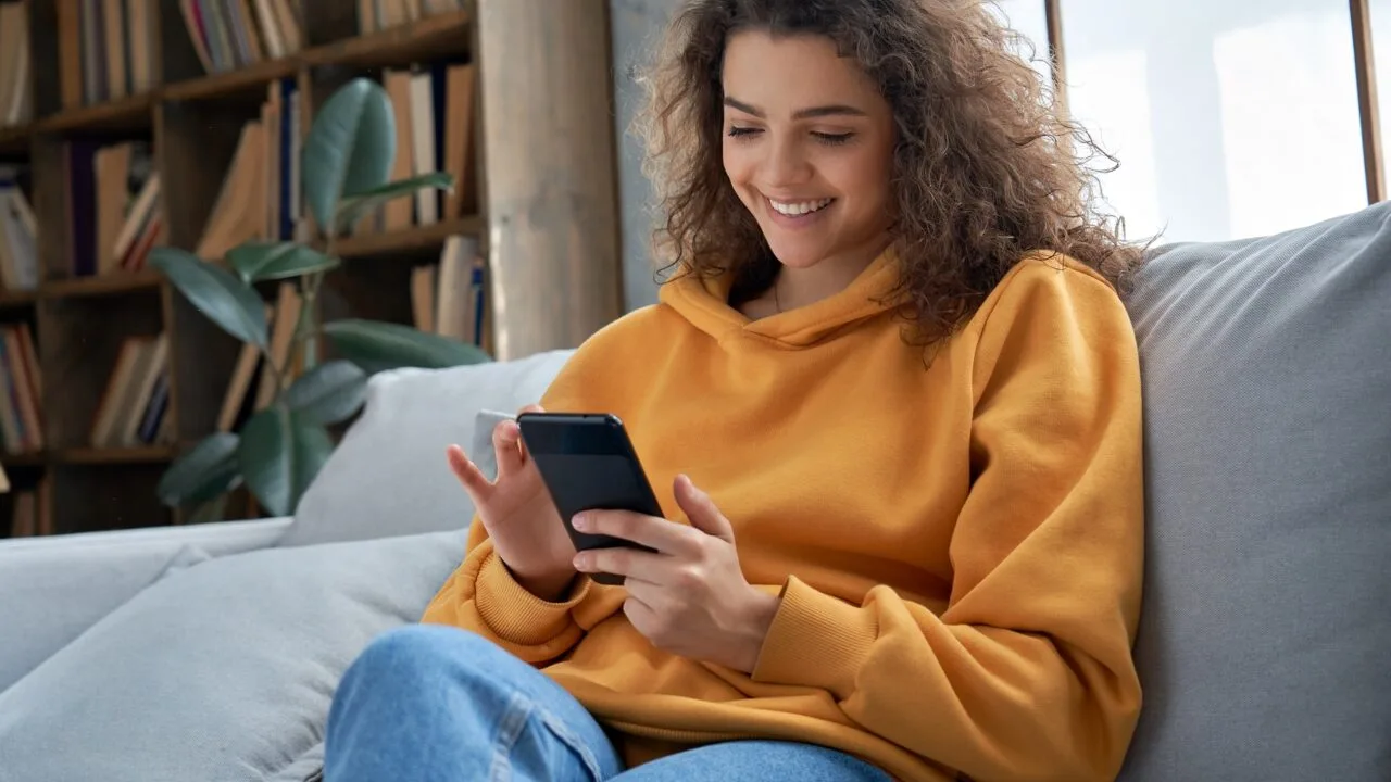 Imagem mostra mulher branca de cabelos castanhos e cacheados, vestindo blusa de frio com capuz amarelo ocre e sorrindo ao mexer no celular, sentada em um sofá branco, no que parece uma sala de estar. Ao fundo, vemos uma estante de livros à esquerda, com um vaso de planta na frente, e uma janela grande ao lado direito, logo atrás do sofá.