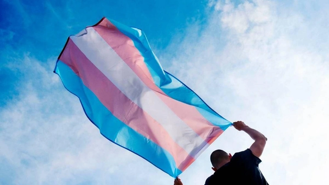 Imagem de uma pessoa segurando a bandeira transgênero contra um céu azul. A bandeira é composta por faixas rosa, azul e branca, simbolizando a luta e a visibilidade da comunidade trans.