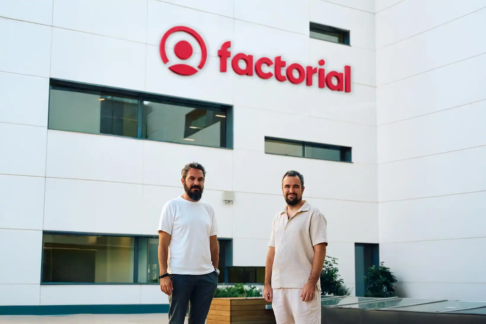Dois homens sorrindo, Jordi e Bernat, em frente ao edifício da Factorial, uma empresa de tecnologia, com a logomarca visível na fachada, em um ambiente corporativo moderno.