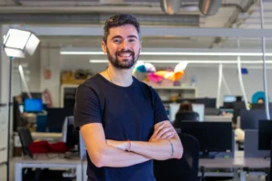 Imagem de homem branco vestindo camiseta preta, sorrindo, de braços cruzados em ambiente corporativo descontraído